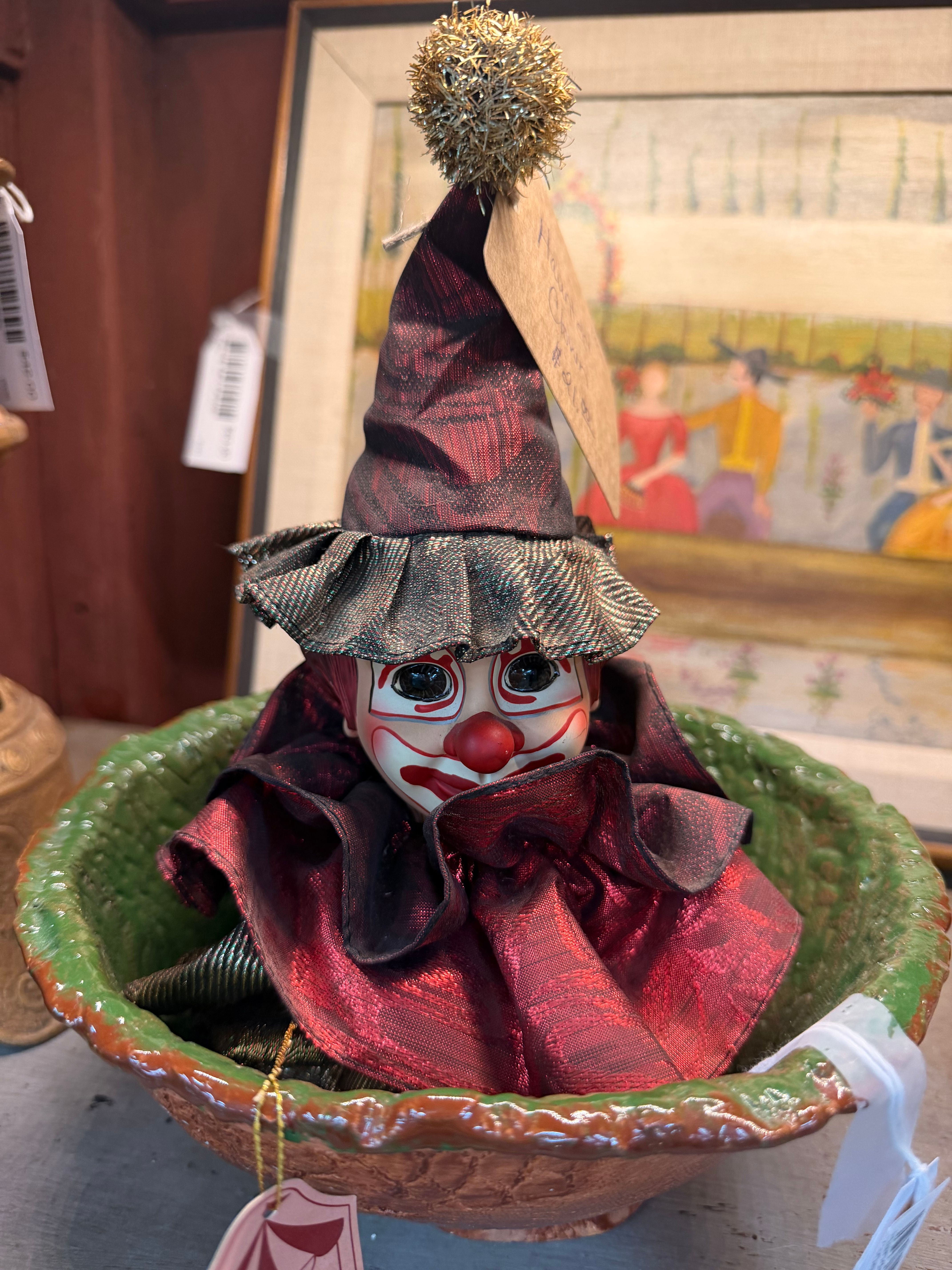 A vintage porcelain clown doll in a red and black silk harlequin costume and pointed hat, resting in a green ceramic bowl