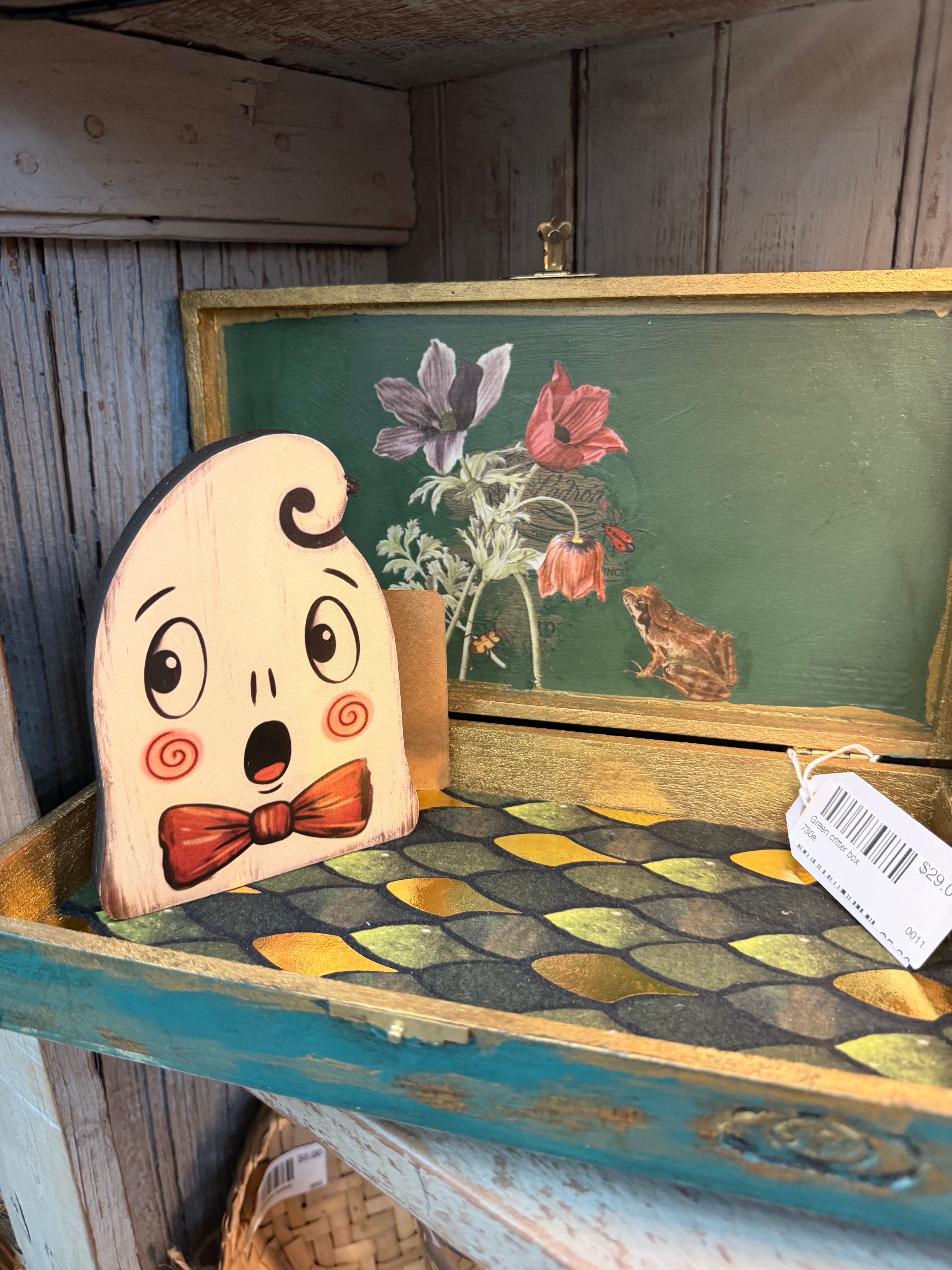 A folk-art wooden ghost cutout with a surprised face and red bow tie, beside a green decoupage tray with florals and a frog