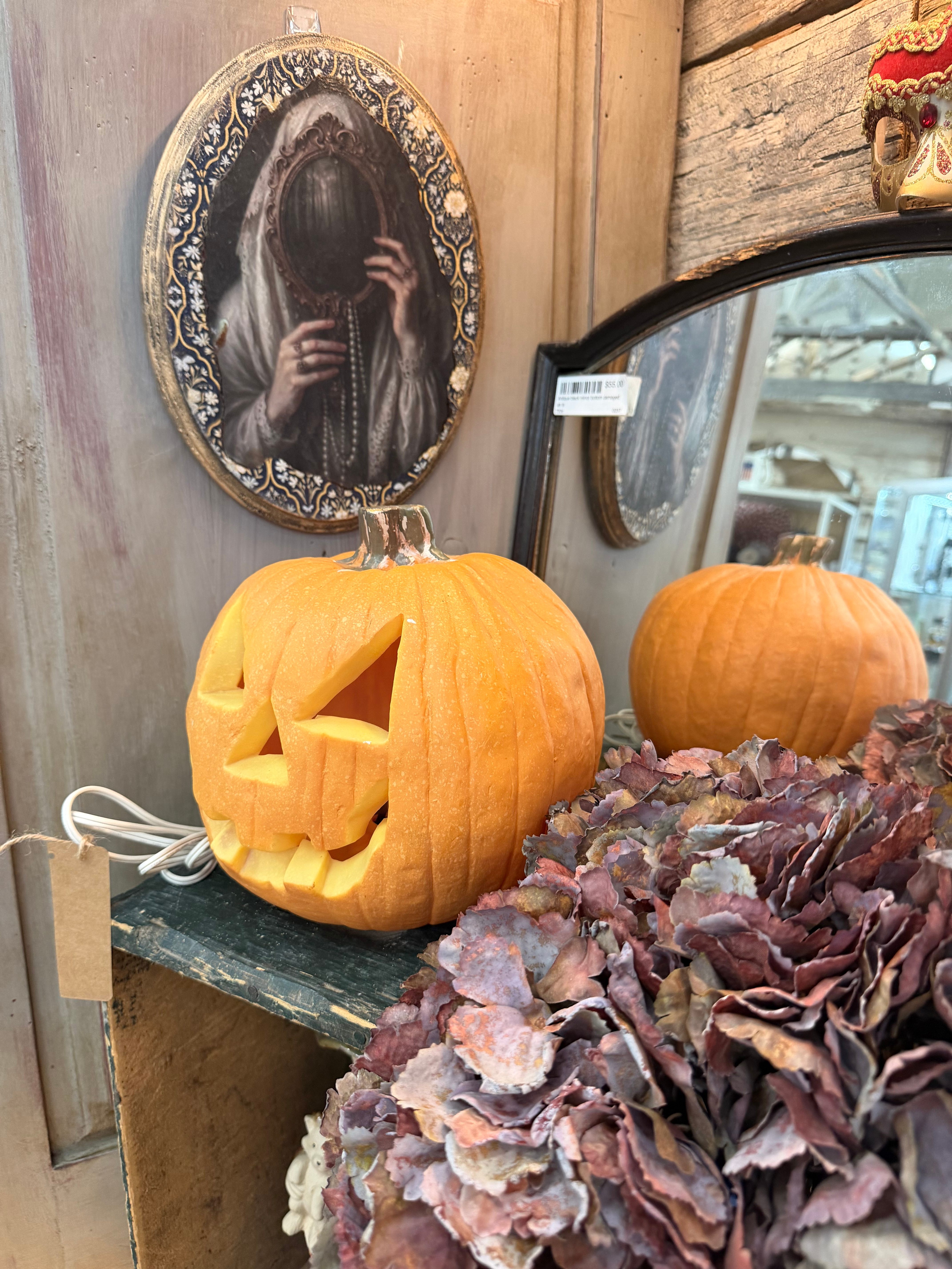 A glowing carved jack-o-lantern on a shelf with a veiled-woman art plaque above and dried hydrangea nearby