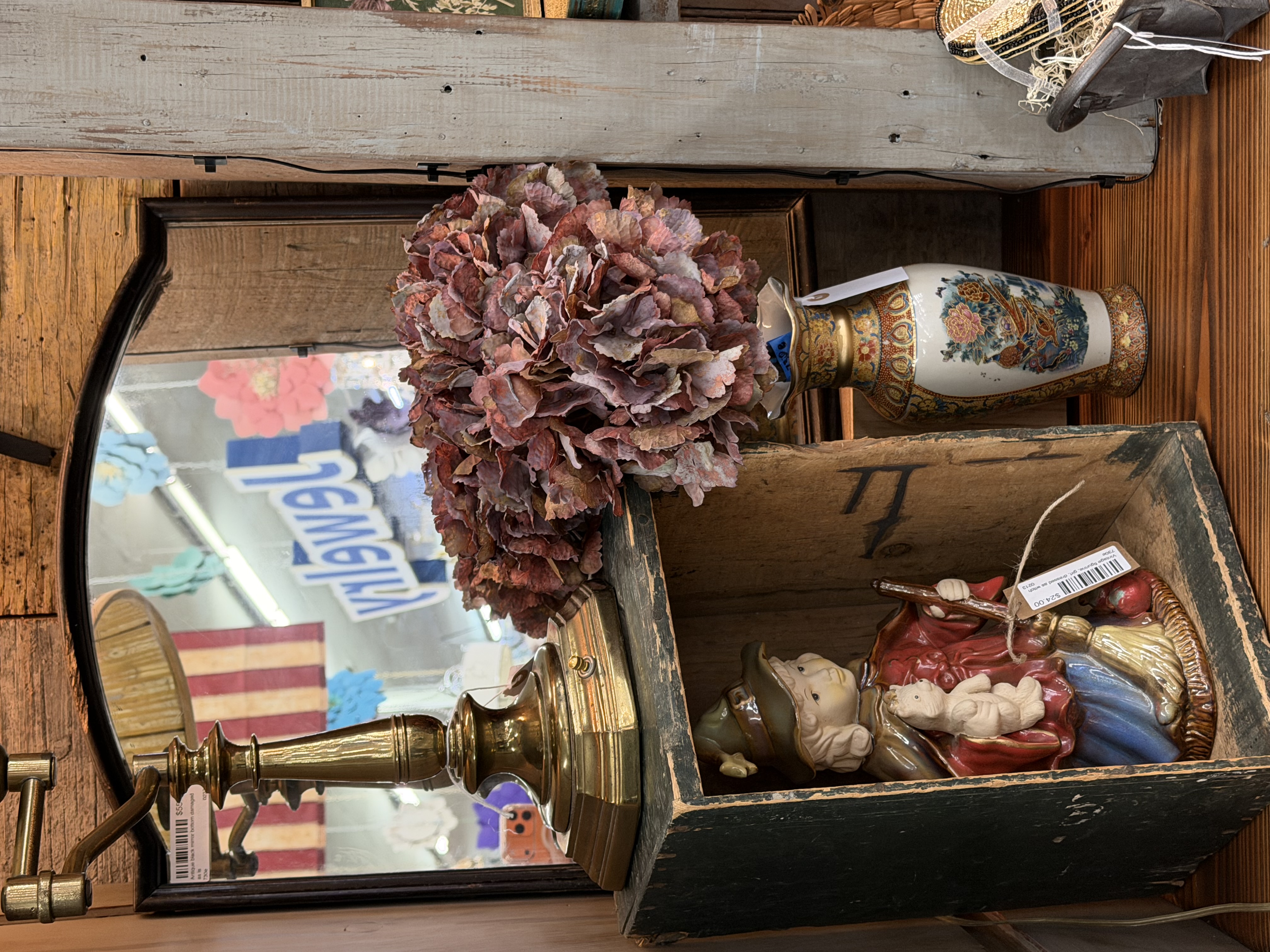 A ceramic figurine of a girl in a witch hat holding a cat, displayed in a dark crate box beside a brass lamp and dried burgundy hydrangea
