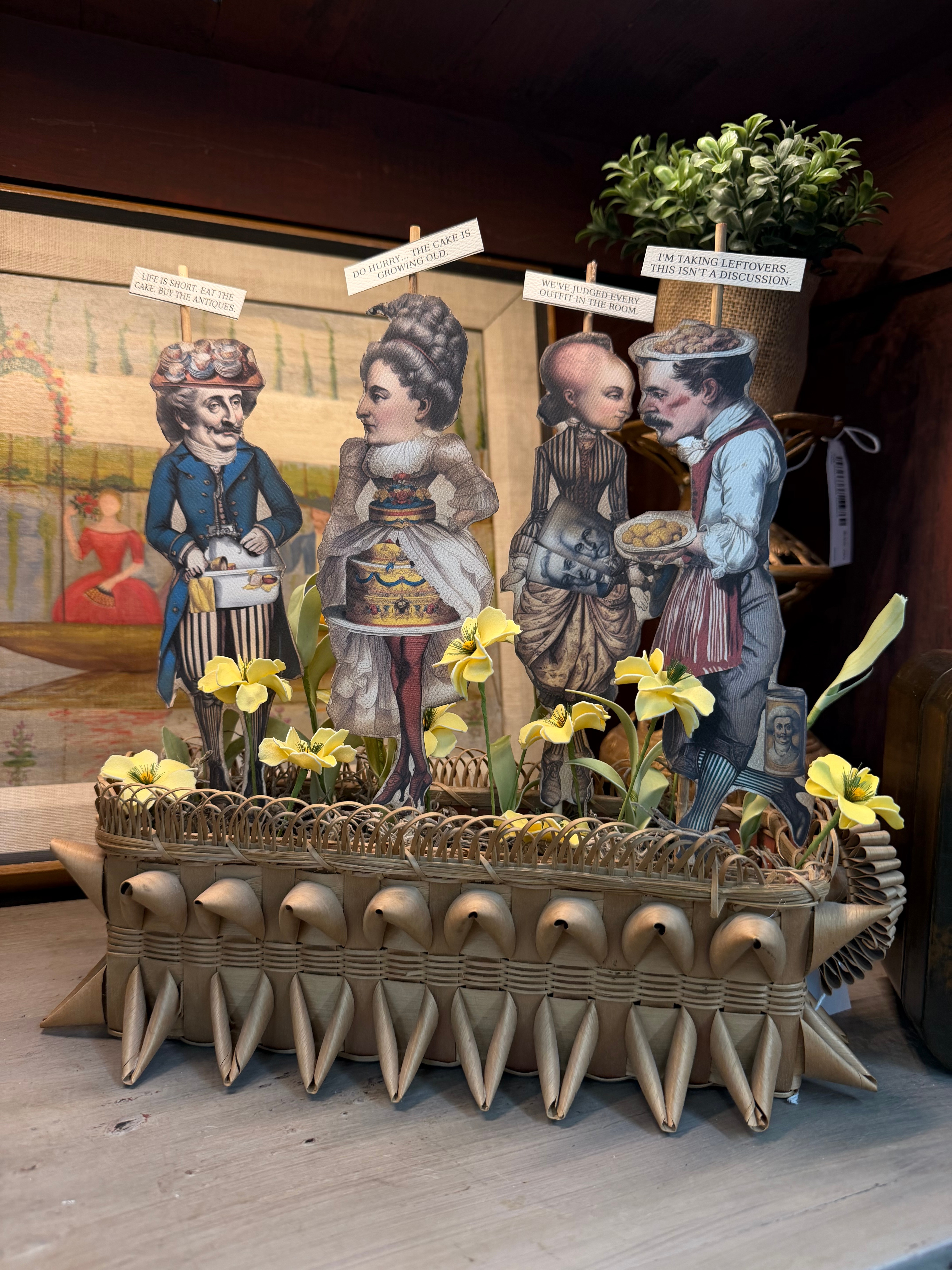A whimsical centerpiece of Victorian paper dolls with witty sayings in a woven basket surrounded by yellow blooms